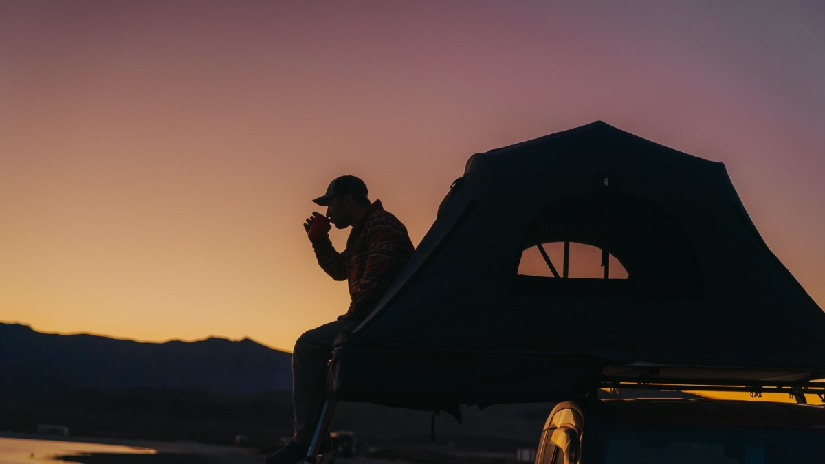 Silhouette af en mand, der sidder på kanten af sit tagtelt og drikker noget