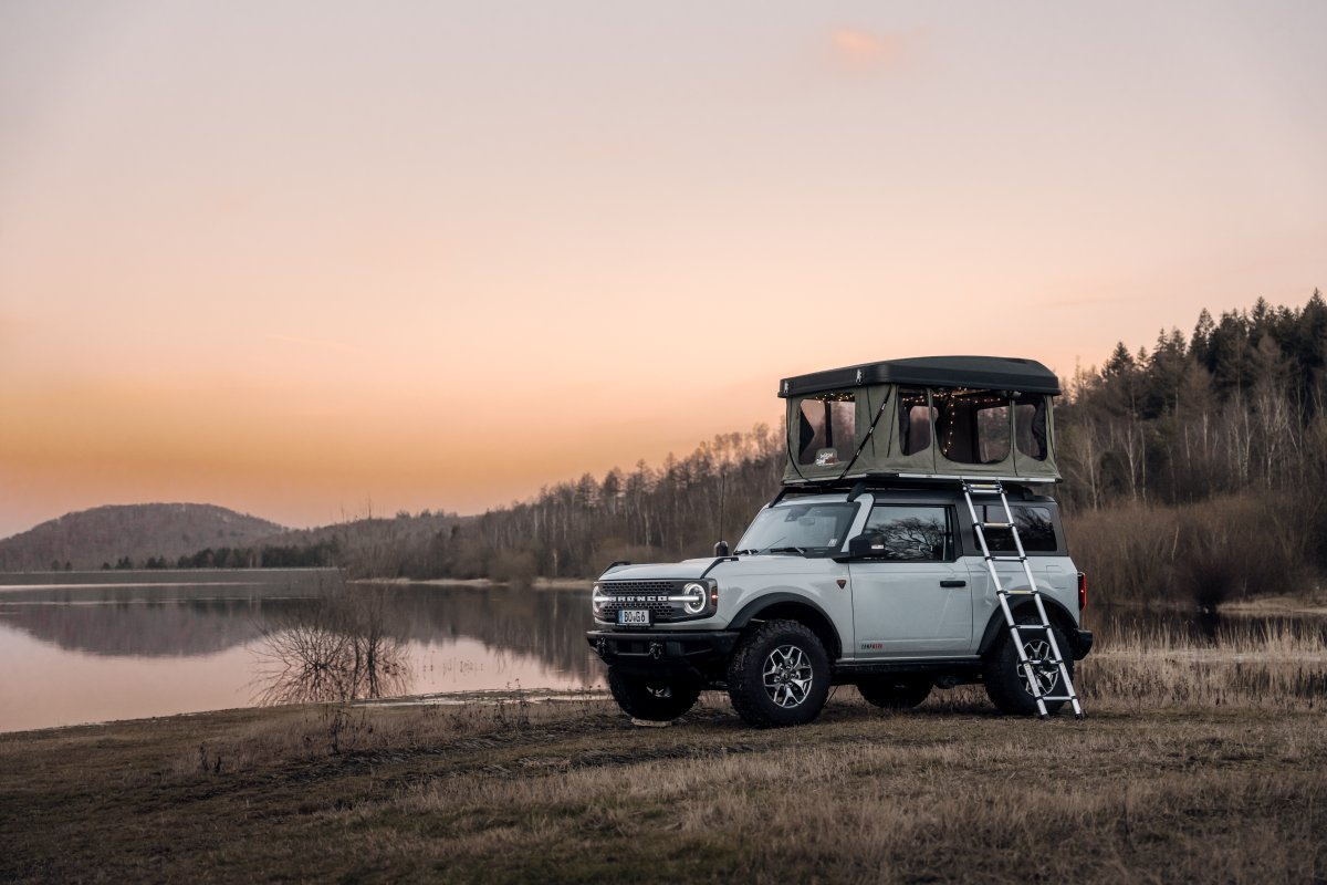 Bil med hard shell-tagtelt til 1 person parkerer ved solnedgang ved en sø